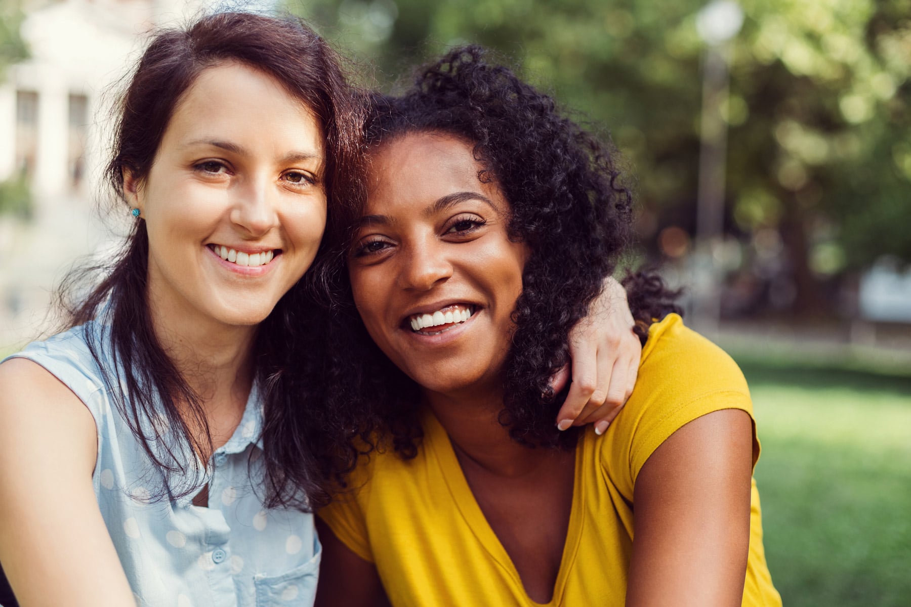 smiling white woman embracing and wrapping arm around smiling black woman