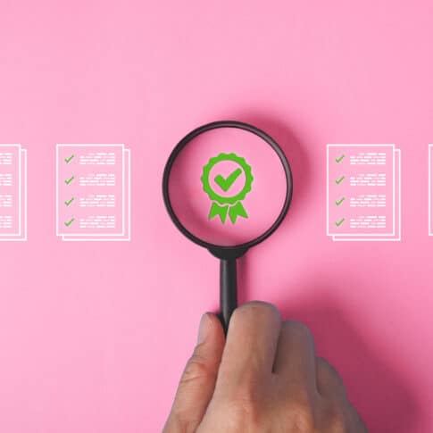 A hand holding a magnifying glass over a pink background with a green certificate check mark and documents in quality assurance concept.