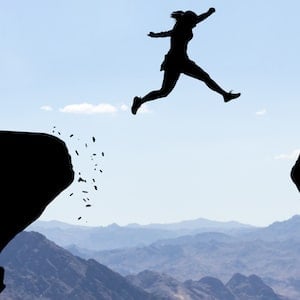 Woman jumping over abyss in front of mountain background.