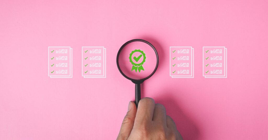 A hand holding a magnifying glass over a pink background with a green certificate check mark and documents in quality assurance concept.