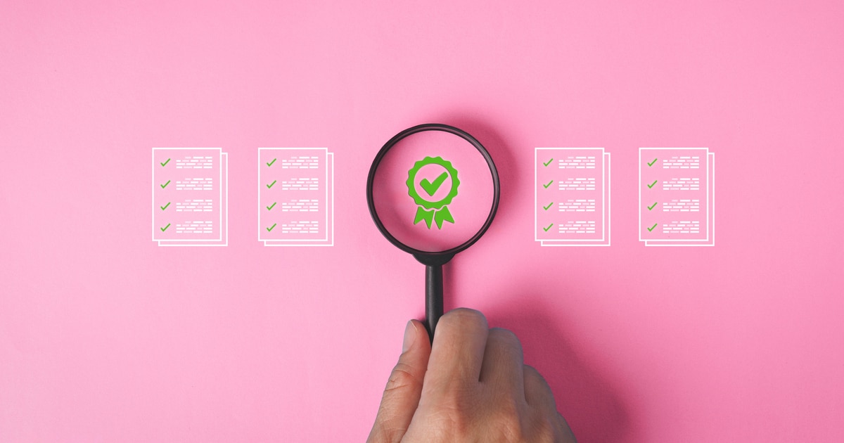 A hand holding a magnifying glass over a pink background with a green certificate check mark and documents in quality assurance concept.